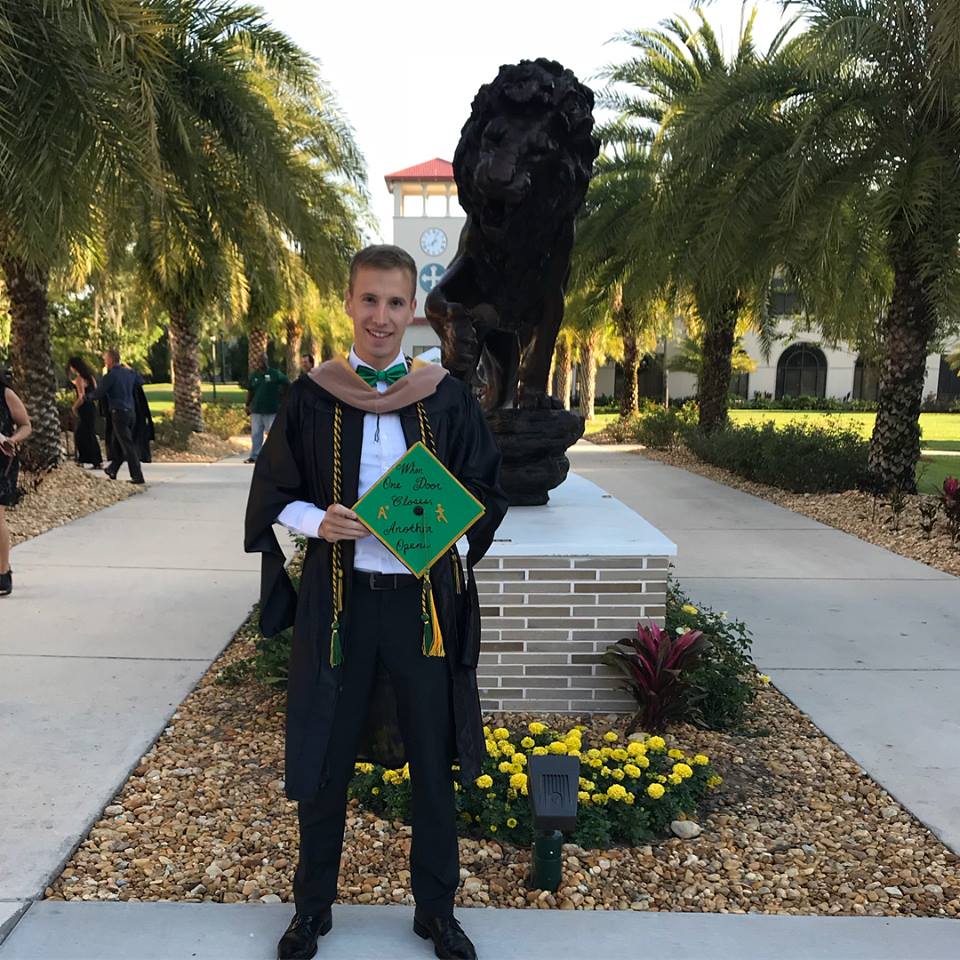 Saint Leo student Niclas Bez posing next to a copper lion statue on University Campus in his cap and gown as he was able to walk at graduation in April 2018 prior to formally completing his MBA coursework in August 2018